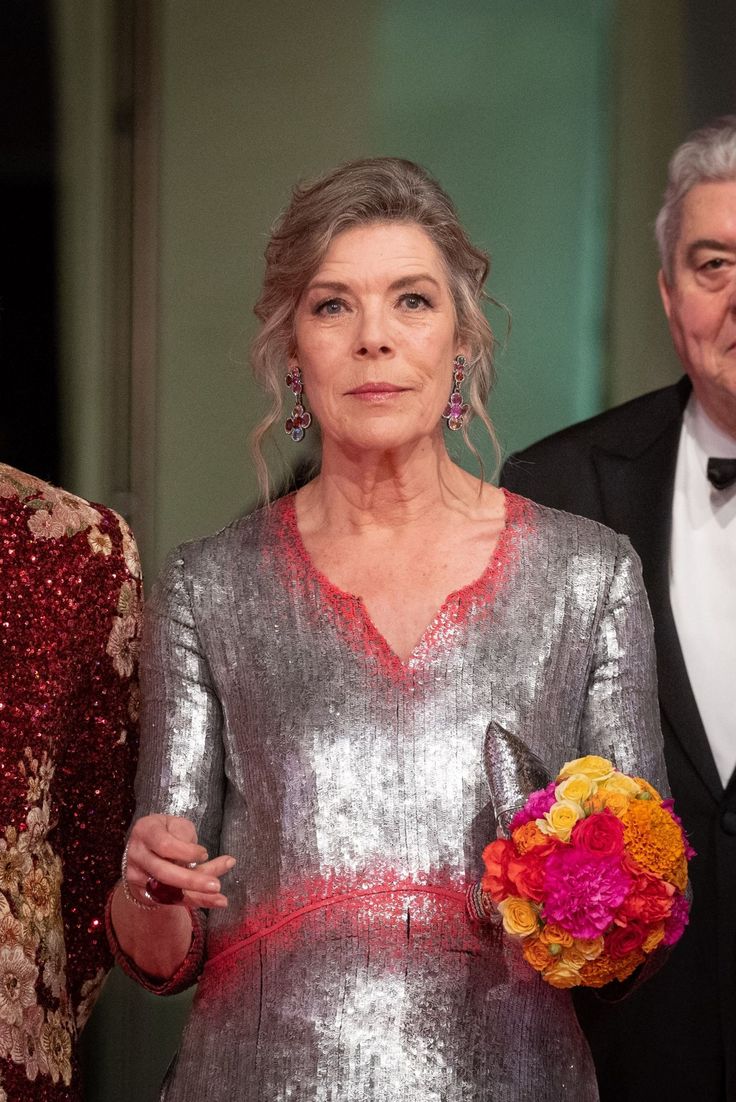 an older woman wearing a silver dress and holding a flower in her hand while standing next to two older men