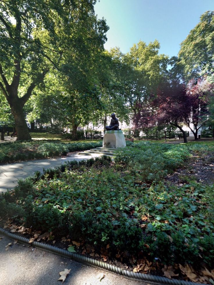 a statue in the middle of a park surrounded by trees and bushes on a sunny day