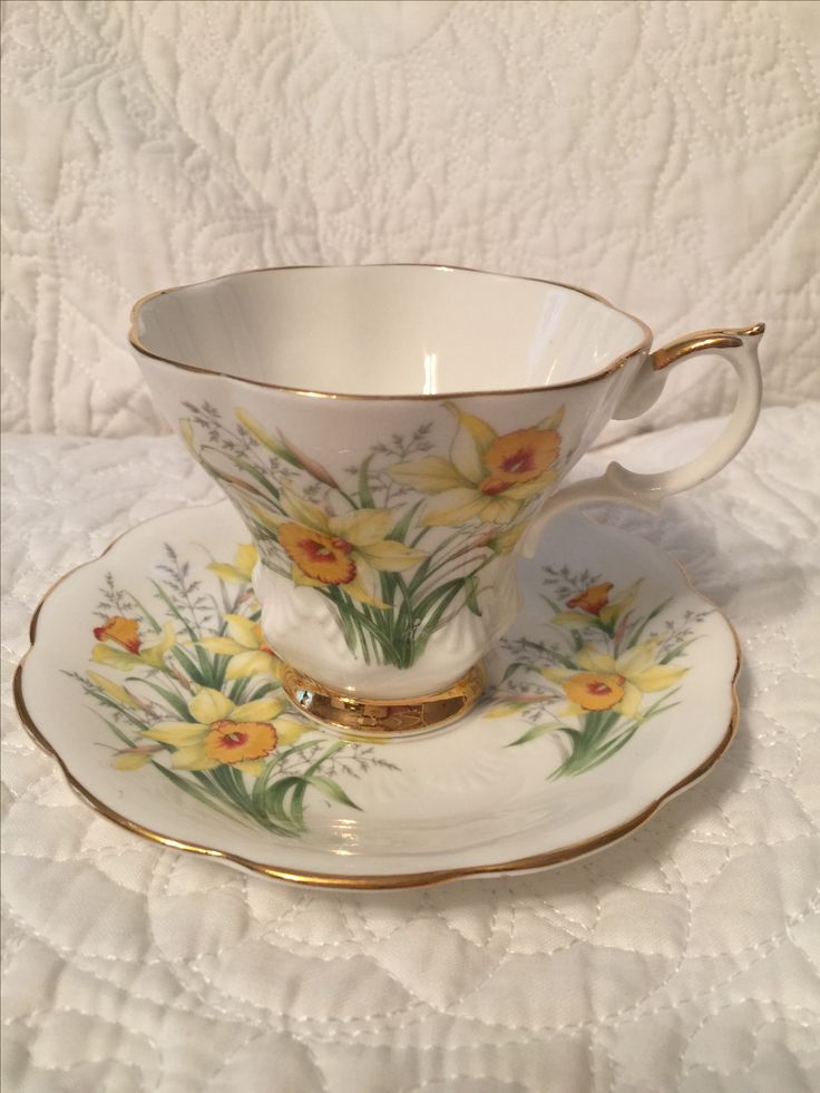 a tea cup and saucer with yellow flowers on the side, sitting on a white tablecloth