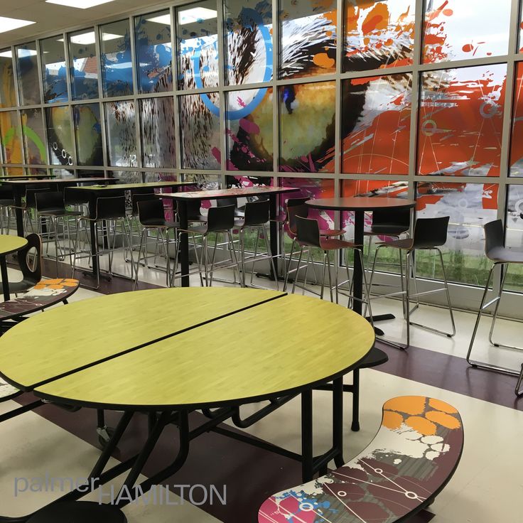 an empty cafeteria with tables and skateboards in front of large windows that have colorful paintings on them