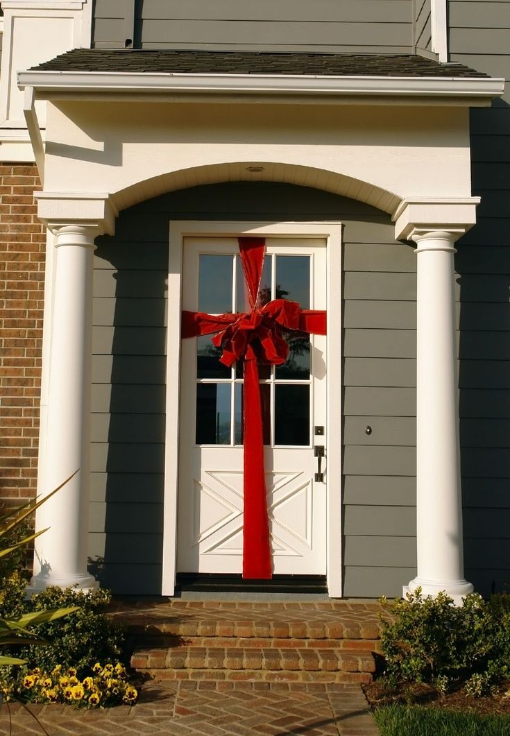 a large red bow on the front door of a gray house with white pillars and windows