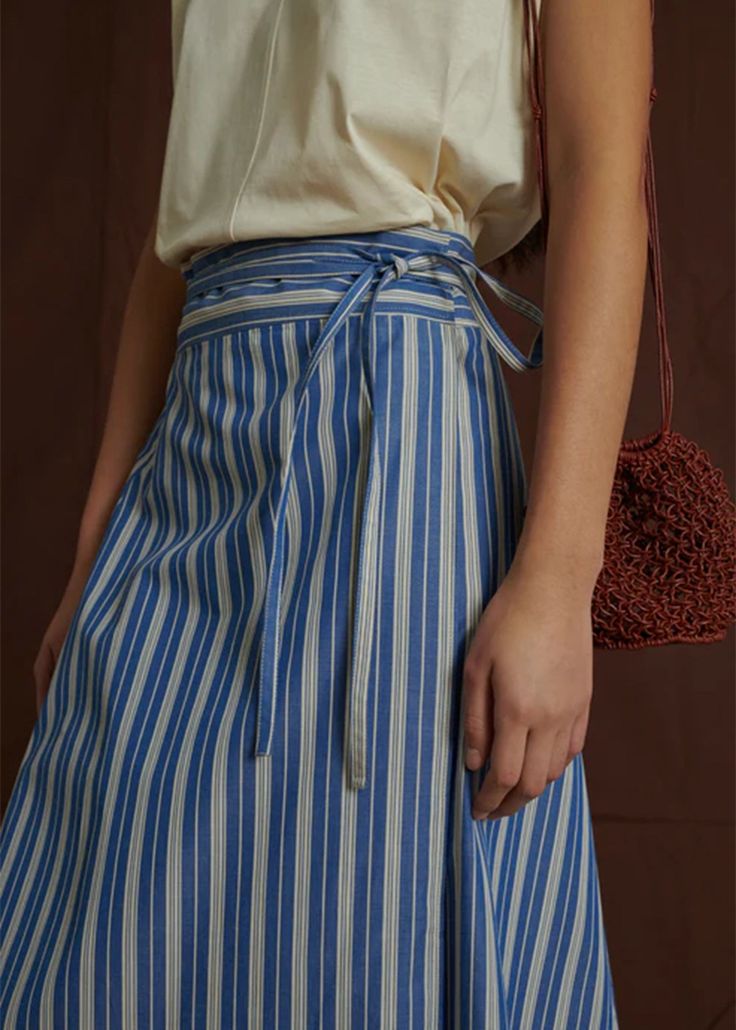 Soeur's Reine Skirt is a mid-length wrap skirt made from a emerized cotton poplin for a softer, more flexible feel. The Reine Skirt features a trapeze silhouette, a wide belt that ties at the waist and allows for an adjustable fit, and a blue and white pinstripe print throughout. 100% cotton machine wash cold, tumble dry low Cotton Asymmetrical Skirt For Daywear, Cotton Tie Waist Skirt For Spring, Cotton Skirt With Tie Waist For Spring, Asymmetrical Cotton Wrap Skirt For Workwear, Cotton Maxi Skirt For Summer Workwear, Asymmetrical Tie Waist Relaxed Skirt, Asymmetrical Relaxed Skirt With Tie Waist, Summer Cotton Tiered Wrap Skirt, Summer Daywear Skirt With Tie Waist