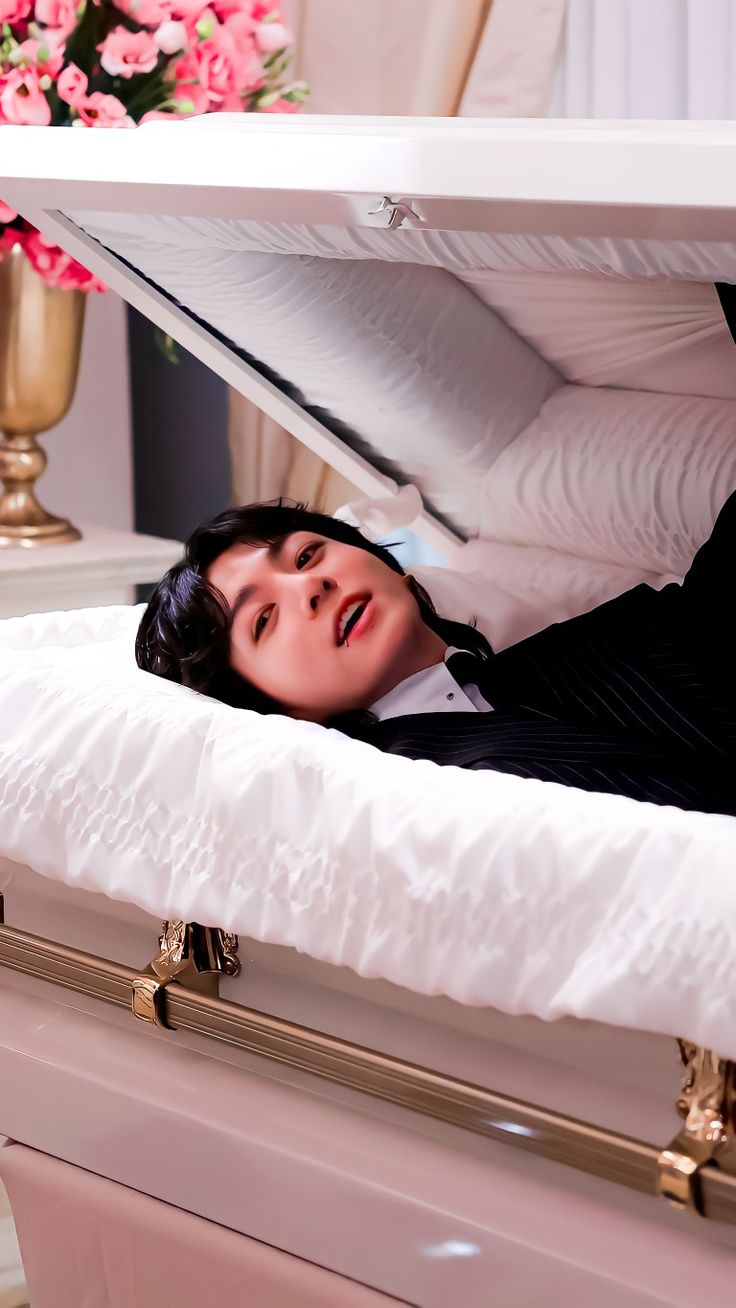 a man in a suit is laying down on a white casket with flowers behind him