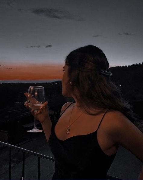 a woman holding a glass of wine on top of a balcony at sunset with mountains in the background