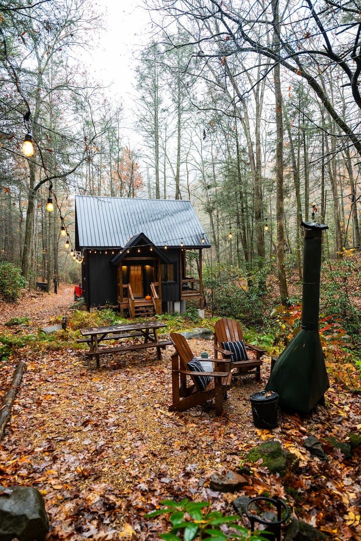 a small cabin in the woods with picnic tables and chairs next to it, surrounded by trees