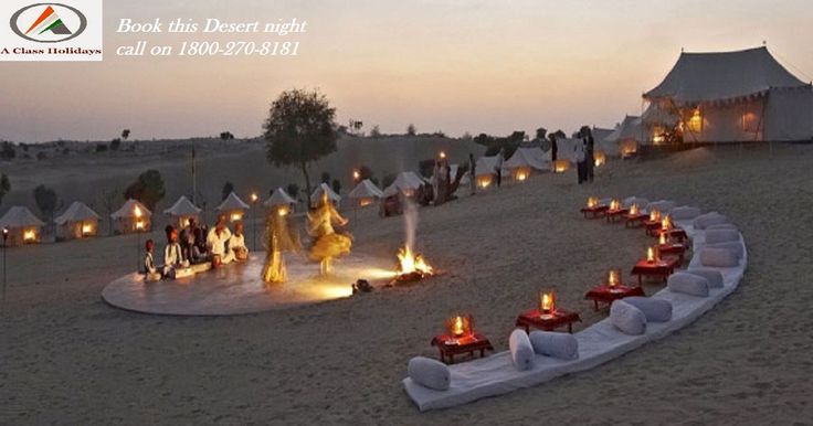 a group of people standing around a fire pit on top of a sandy field next to tents