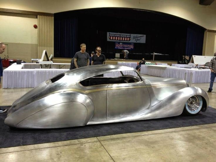 two men standing next to a silver car on display in a building with people looking at it