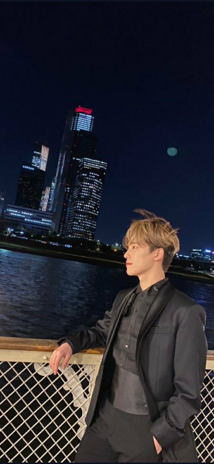 a young man standing on top of a bridge next to the water at night with city lights in the background