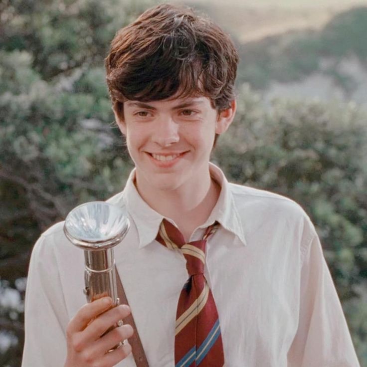 a young man wearing a tie and holding a silver object in his hand with trees in the background