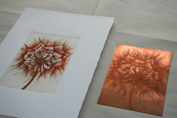 two paintings are displayed on a table with white paper and red ink, one has a dandelion in the middle