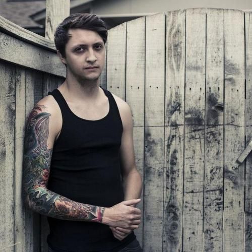 a man with tattoos standing next to a wooden fence and holding his hands on his hips