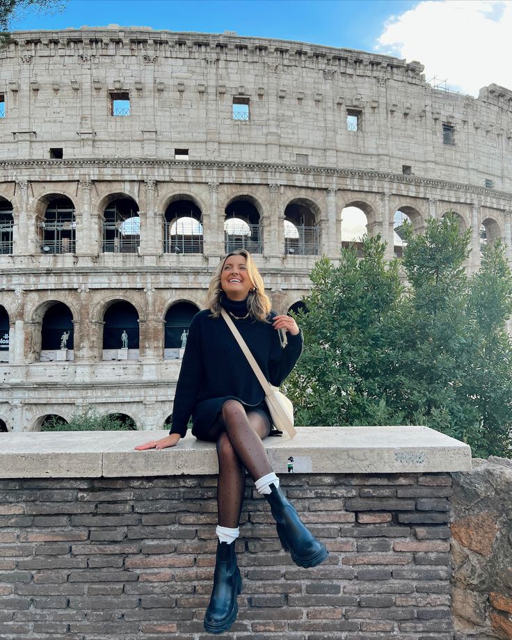 a woman sitting on top of a brick wall in front of an ancient arena building