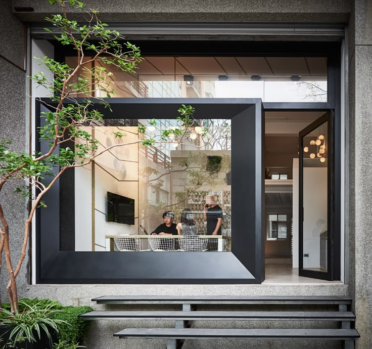 two people sitting at a table in front of a large window with plants on it