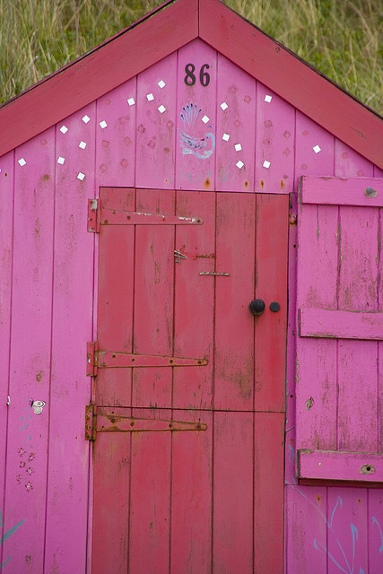 a pink building with a red door and window on the outside, in front of some tall grass