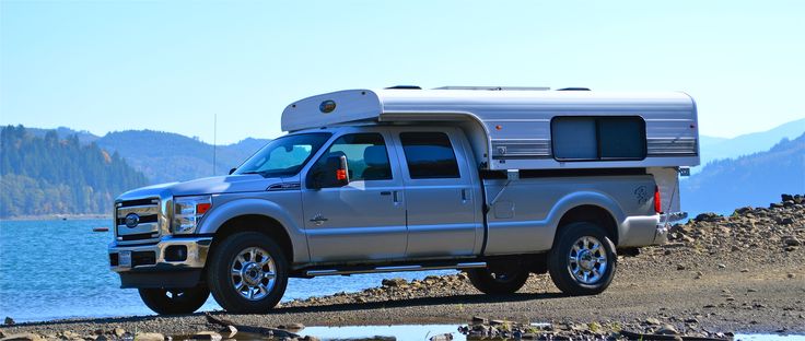 a silver truck with a camper on the back parked near water and mountains in the background