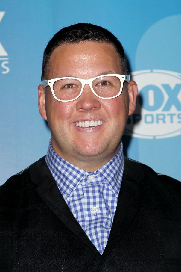 a man wearing glasses and a blue shirt smiles at the camera while standing in front of a fox sports logo