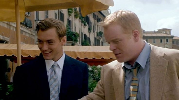 two men standing next to each other in suits and ties looking at something on the table