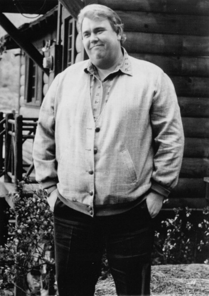 a man standing in front of a log cabin with his hands in his pockets and looking at the camera