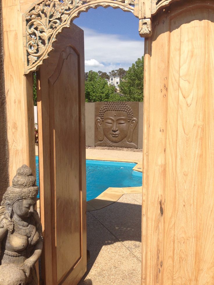 an open wooden door leading to a pool with buddha statues in the foreground and a carved wood doorway