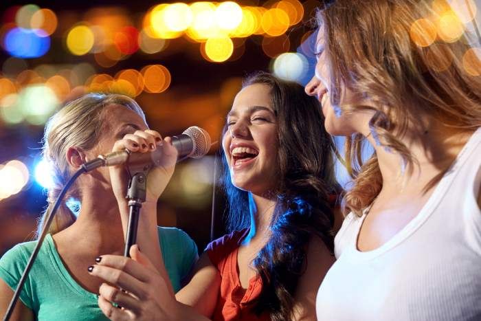 three women singing into microphones in front of blurry city lights at night time