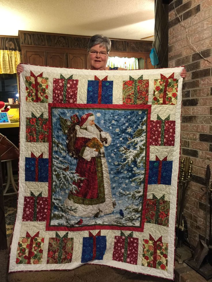 a woman holding up a christmas quilt in front of a fireplace