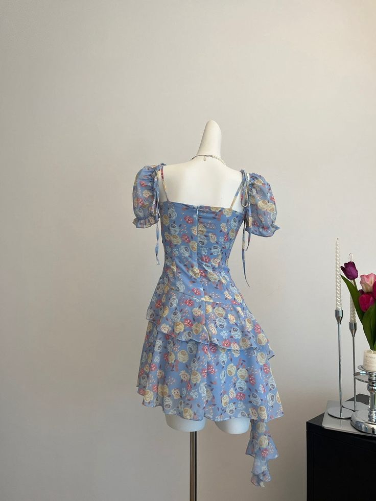 Circuitous and crafted with care, this blue floral dress weaves its way through timeless fashion with a modern twist. The layers of lightweight fabric create a gentle cascade, giving this dress a buoyant, ethereal quality that’s perfect for spring and summer events. Decorated with a vibrant array of floral patterns in soft hues, the dress features dainty shoulder straps and a rosette detail for a touch of romance. Its asymmetrical hemline and flowing silhouette ensure it moves gracefully with th Spring Floral Print Mini Tiered Dress, Feminine Summer Tiered Dress For Garden Party, Feminine Tiered Summer Dress For Garden Party, Feminine Tiered Dress For Summer Garden Party, Summer Floral Dress With Square Neck And Ruffles, Light Blue Sleeveless Floral Dress For Summer, Spring Square Neck Feminine Tiered Dress, Blue Chiffon Dress With Square Neck, Blue Chiffon Square Neck Dress