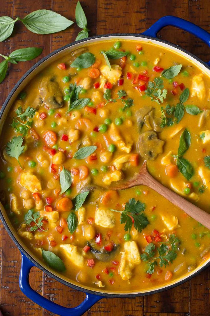 a blue pot filled with yellow curry and vegetables on top of a wooden table next to a green leafy garnish