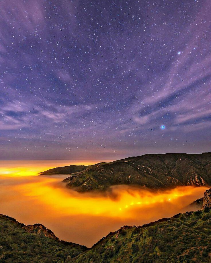 the night sky with clouds and stars over mountains in the foreground is lit by bright lights