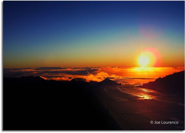 the sun is setting over some mountains with clouds in the foreground and water on the ground below