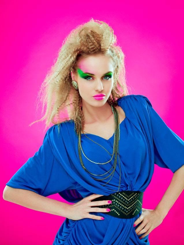 a woman with bright makeup and green eyeshades posing for a photo in front of a pink background