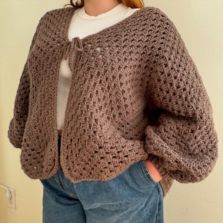 a woman wearing a brown crochet sweater and jeans standing in front of a wall