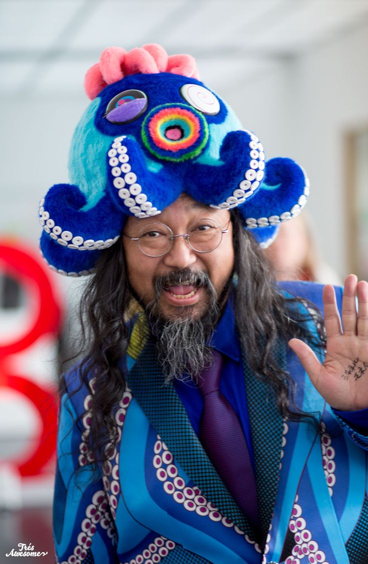 a man with long hair wearing a blue hat and purple tie holding his hand up