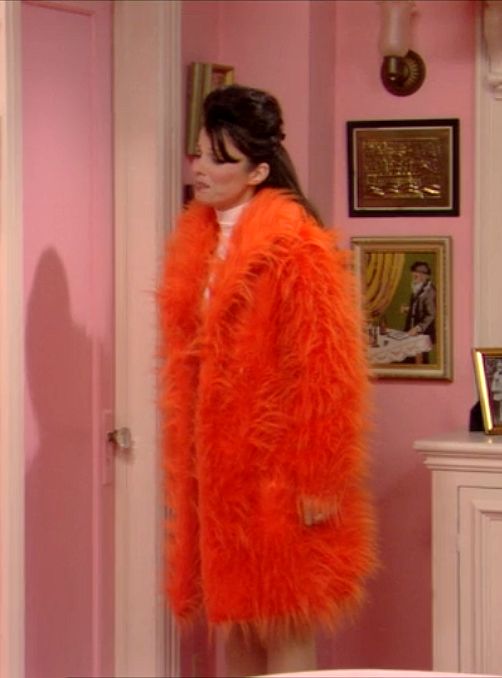 a woman in an orange fur coat standing in a pink room