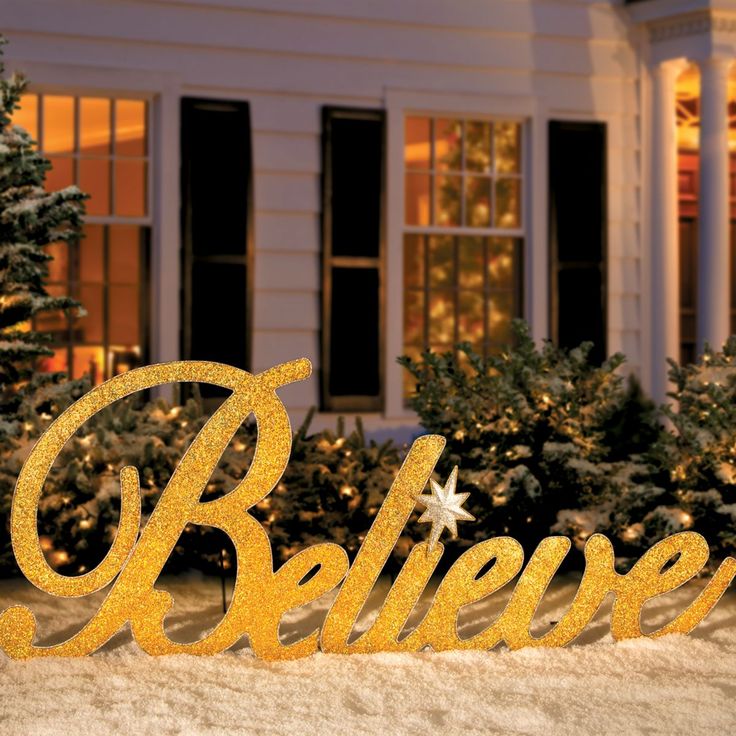 a gold believe sign in front of a white house with christmas trees and evergreens