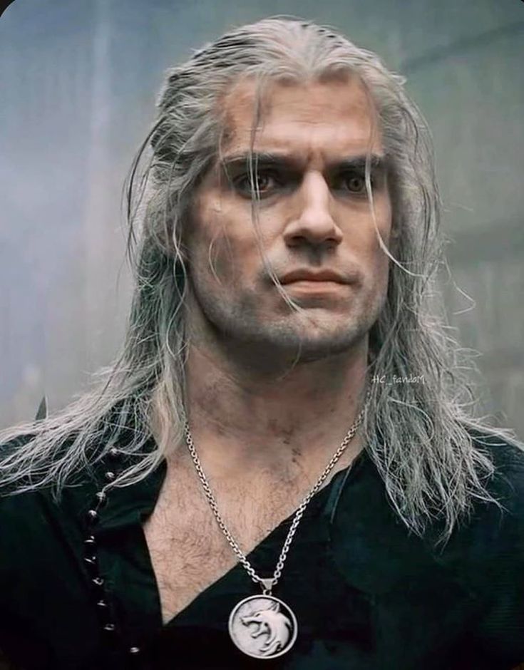 a man with long white hair wearing a black shirt and silver necklace, looking at the camera