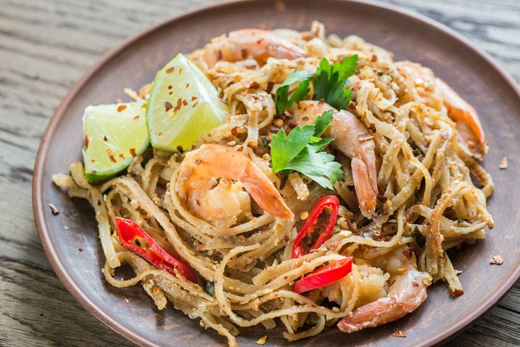 a plate full of noodles with shrimp, limes and cilantro on the side