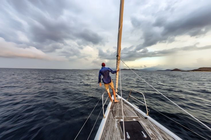 a man standing on the bow of a sailboat looking out at an island in the distance