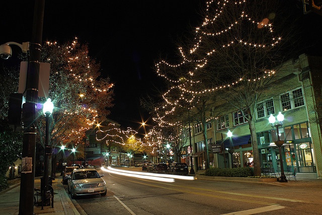 a city street at night with christmas lights on trees and cars driving down the road