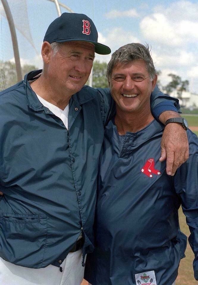 two men standing next to each other in front of a baseball field with one man hugging the other