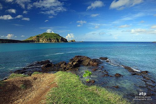 an island in the middle of the ocean with blue sky and clouds above it is surrounded by green grass