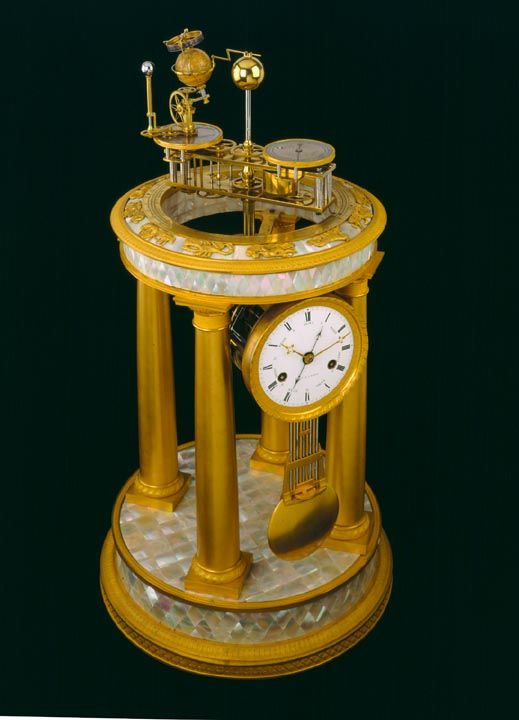 a gold clock sitting on top of a marble table next to a black background with columns