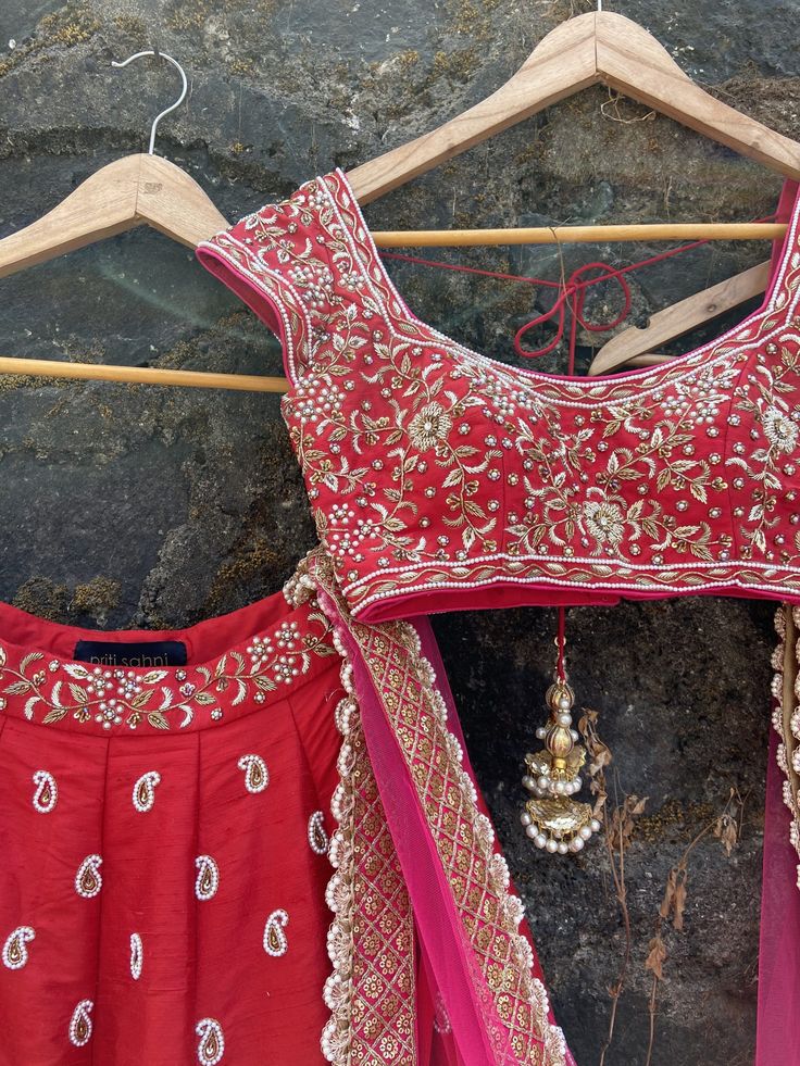 This bridal red raw silk lehenga is embellished with all over paisley pearl and zardozi work with a pink sari sequin wool border. It is paired with a  red raw silk blouse with zardozi, pearl hand embroidery all over with pink accents. This bridal beauty is completed with a pink net dupatta with button work all over and a scalloped border. Traditional Embellished Pre-draped Saree For Festive Season, Red Pre-draped Saree For Eid Wedding, Red Resham Embroidery Pre-draped Saree For Wedding, Traditional Embellished Pre-draped Saree In Raw Silk, Red Silk Sharara With Dori Work, Red Embellished Sharara For Navratri, Red Pre-draped Saree For Reception At Eid, Red Silk Anarkali Set With Dori Work, Red Embellished Saree For Reception