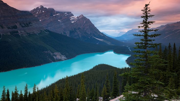 a beautiful blue lake surrounded by mountains under a cloudy sky in the distance is pine trees