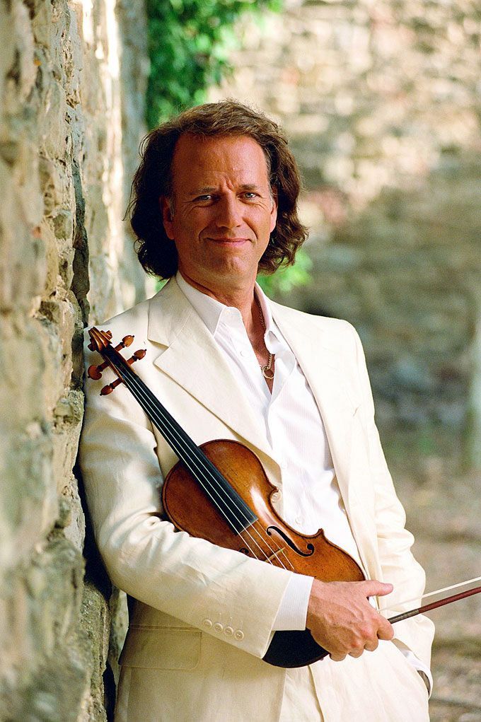 a man wearing a white suit and holding a violin leaning against a stone wall with trees in the background