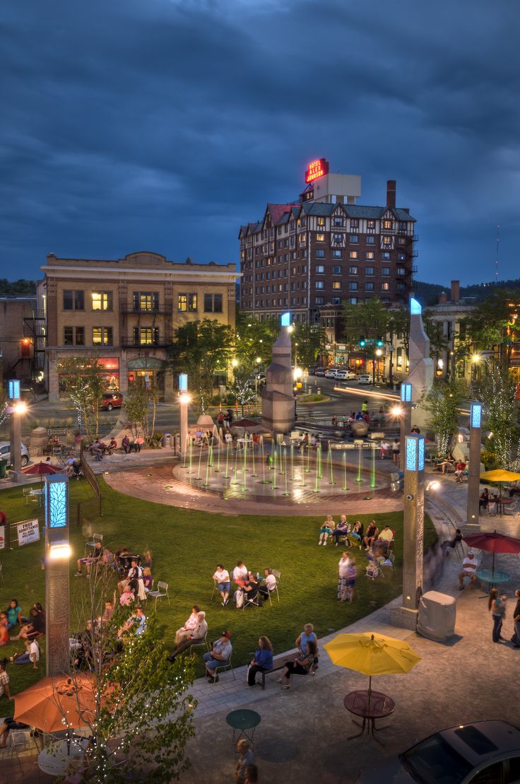 people sitting and walking around in the evening at an outdoor park with fountains, tables, and umbrellas