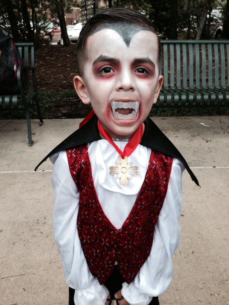 a young boy with his face painted white and wearing a red vest, neck tie and black shoes