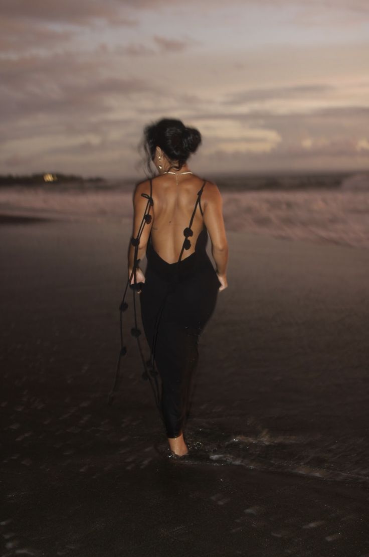 a woman walking on the beach with her back to the camera, wearing a black dress