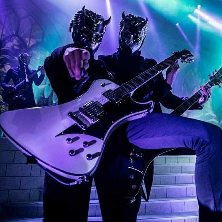 two men in masks are playing guitars on stage with purple lighting and green lights behind them