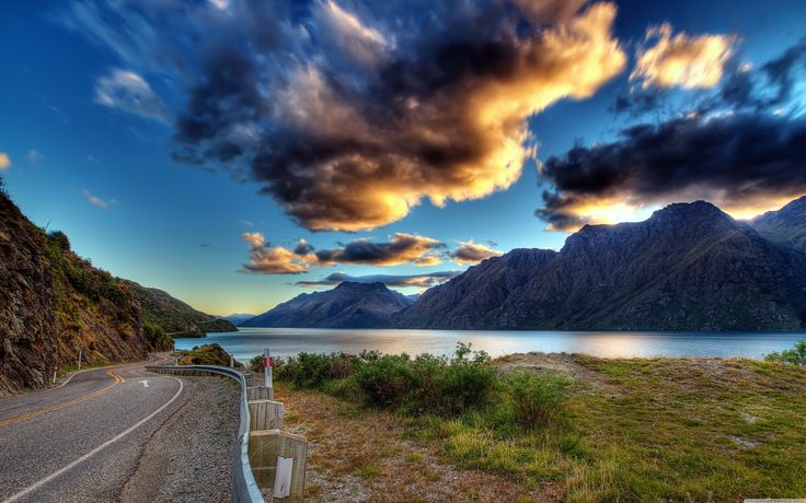 a scenic view of the mountains, water and clouds at sunset or sunrise with an empty road in front of it