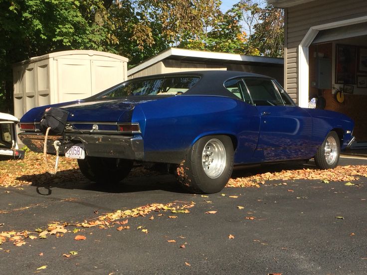 an old blue car parked in front of a garage with autumn leaves on the ground